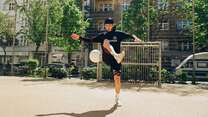 Ein junger Mann, der auf einem städtischen Fußballplatz in sportlicher Kleidung jongliert und dabei einen Fußball in der Luft hält.