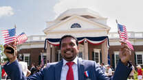 A man smiles while waving an American flag in each hand at a naturalization ceremony.