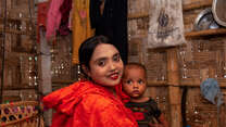 A mother, wearing a bright orange shirt, holds her young child and poses for a photo inside their home in Bangladesh.