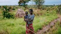 A mother walks through a field while holding her child in her arm.