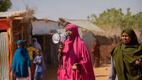 Woman in pink head scarf on a speaker phone, people walking around her
