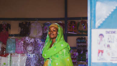 A woman, wearing a vibrant green headscarf and yellow dress, smiles in a shop in Cameroon.
