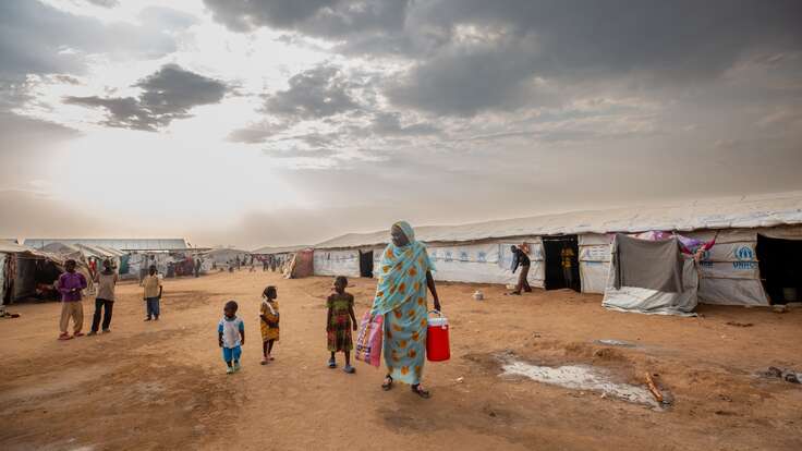 Eine Mutter geht durch eine Notunterkunft für Binnenvertriebene in Südsudan. Sie trägt Wasser, während ihre Kinder hinter ihr herlaufen.